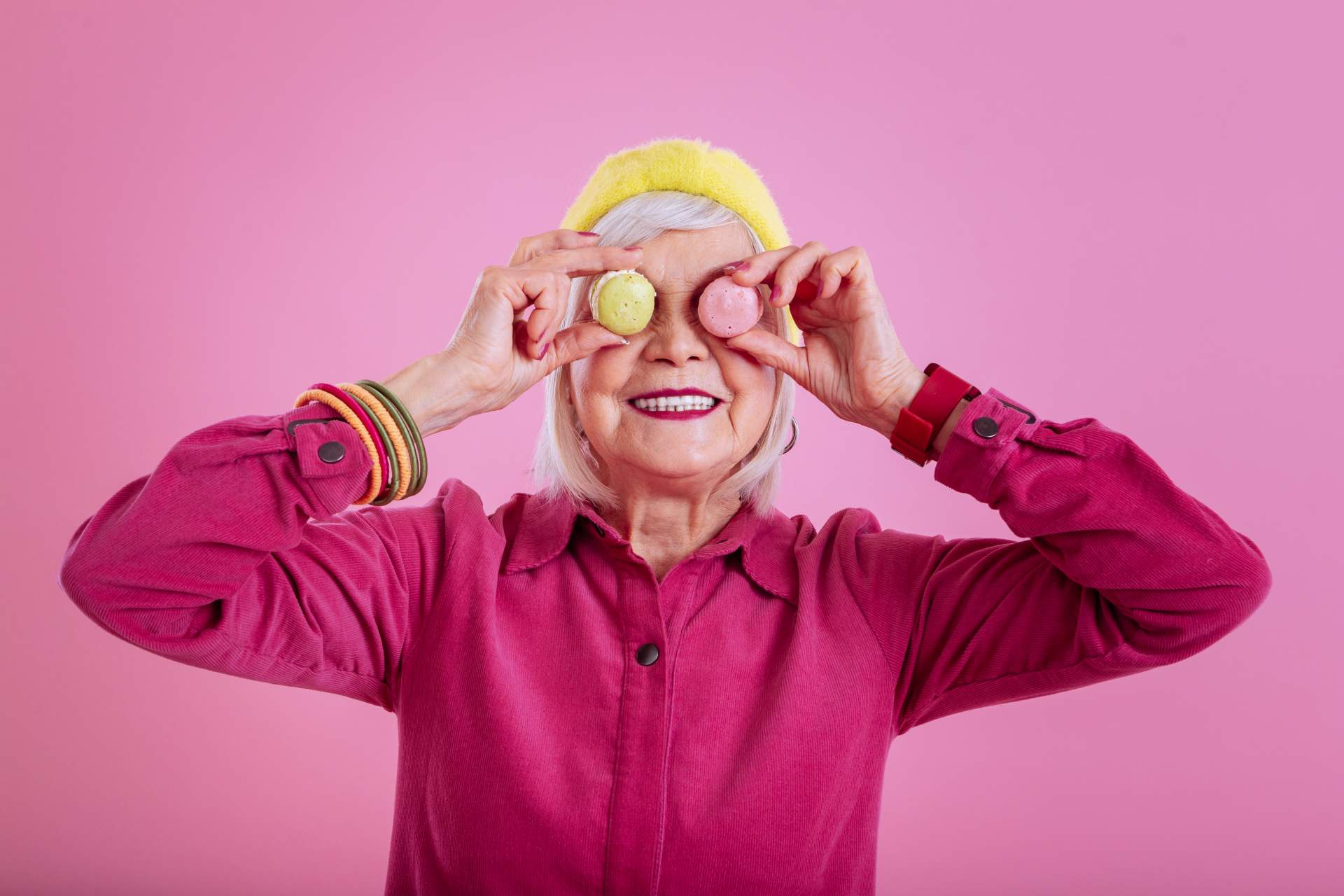 Smiling senior woman in bright clothing holding colorful macarons to her eyes, showing joy, humor, and positive aging.